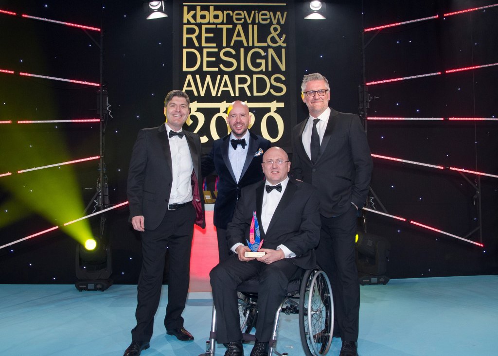 4 men stand on a stage, man using wheelchair is centre place with an award. Background reads 'design awards'